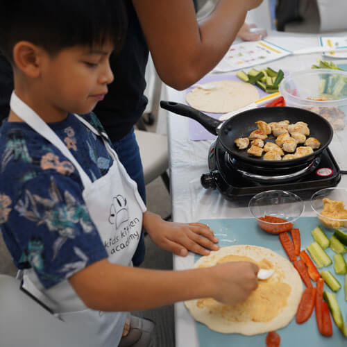 Young dark-haired boy spreading hummus on a pita with chicken grilling on the table